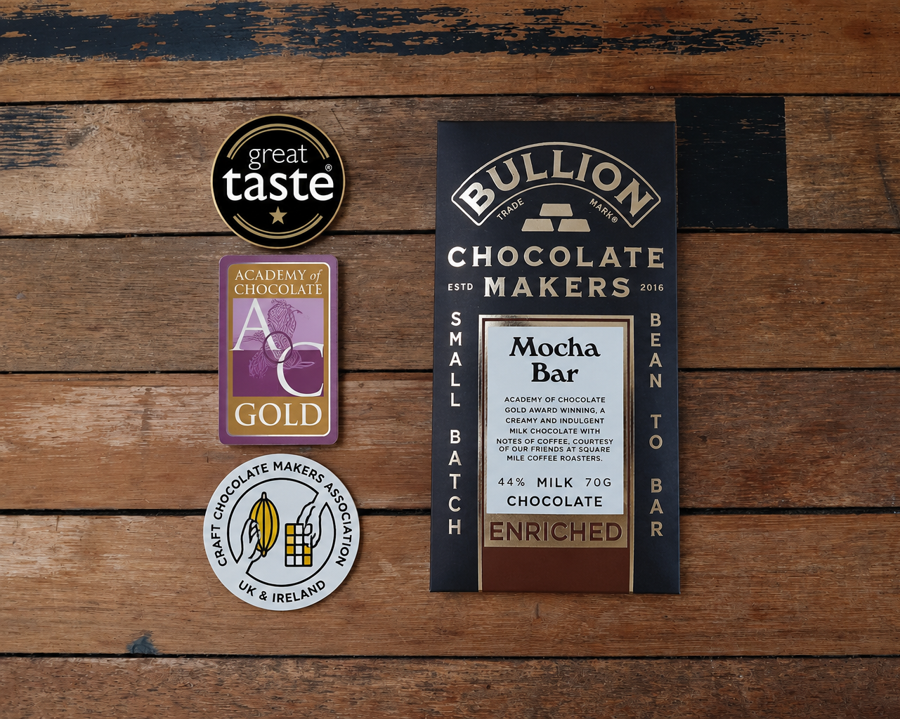 A Bullion Chocolate Makers Mocha Bar is displayed on a rustic wooden background alongside three award badges: Great Taste, Academy of Chocolate Gold, and Chocolate Makers Association UK & Ireland.